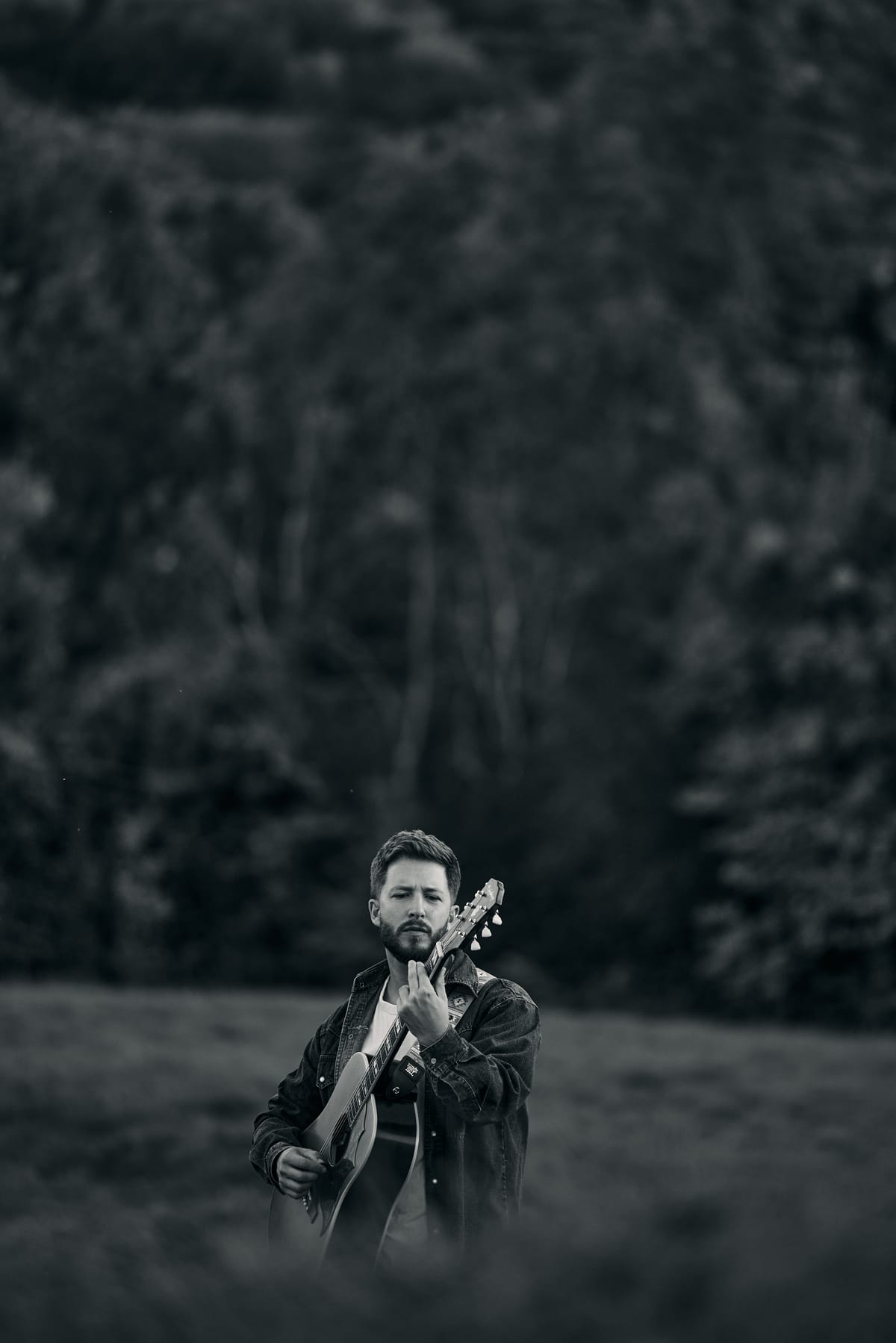 Josh Smith holding his guitar upright in woodland, black and white